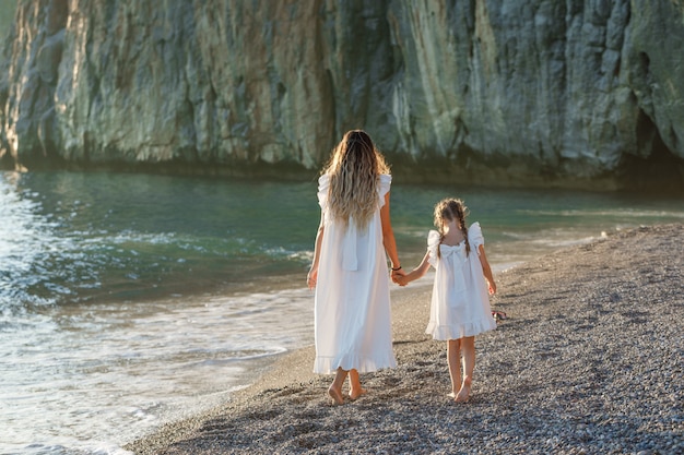 Feliz mãe e filha em vestido branco andando na praia durante o pôr do sol. vista traseira