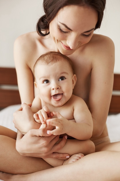 Feliz mãe jovem nua sorrindo abraçando seu bebê recém-nascido agradável, sentado na cama juntos.