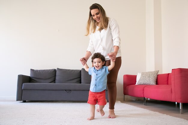 Feliz mãe segurando as mãos do filho e ensinando-o a andar. Menino alegre encaracolado mestiço andando no tapete com os pés descalços com a ajuda da mãe de cabelos compridos. Tempo para a família, infância e conceito do primeiro passo