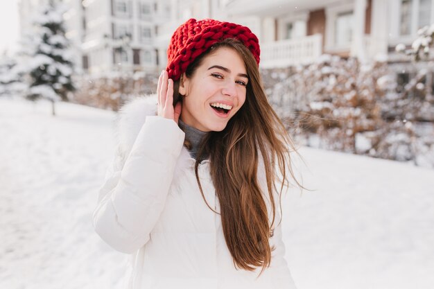 Feliz manhã ensolarada congelada no inverno de uma jovem alegre de chapéu vermelho, com longos cabelos castanhos, se divertindo na rua cheia de neve.