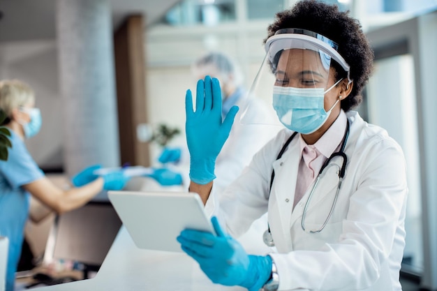 Feliz médico afro-americano acenando durante videochamada enquanto trabalhava no hospital durante a pandemia de coronavírus