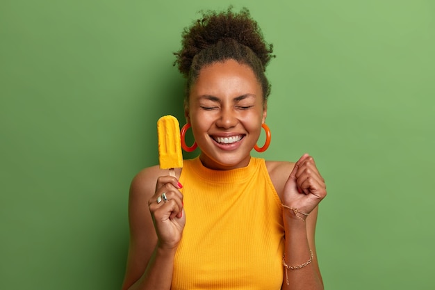 Feliz menina milenar de pele escura come sorvete amarelo com sabor de manga, fecha o punho com a expectativa de que algo incrível aconteça, tem sorriso cheio de dentes, usa roupas de verão brilhantes, isolado no verde