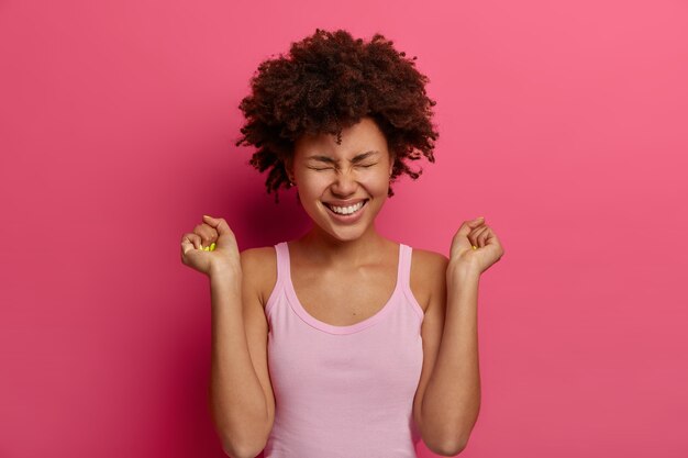 Feliz modelo feminina de pele escura celebra notícias incríveis, levanta os punhos cerrados, sorri amplamente, usa uma camisa casual, posa contra uma parede rosa vibrante, sente-se com sorte e determinada, obteve aprovação