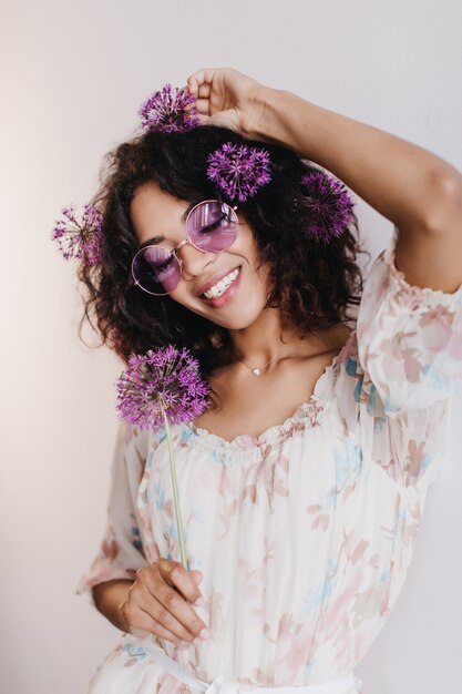 feliz modelo feminino africano com cabelo curto, sorrindo com os olhos fechados. Foto interna de uma garota negra satisfeita posando com flores roxas.