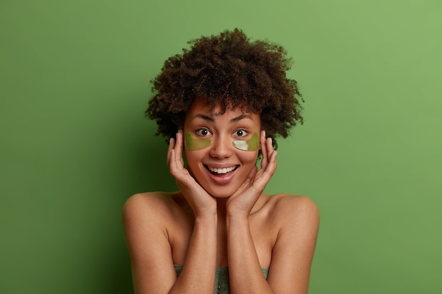 Foto grátis feliz modelo feminino de pele escura com visual fresco, aplica adesivos verdes de hidrogel anti-rugas, toca a pele macia, sorri feliz, modelos em ambientes internos contra paredes vivas e brilhantes. beleza, bem-estar