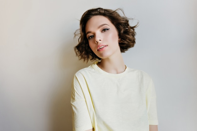 Feliz modelo feminino europeu com cabelo curto escuro, desfrutando da sessão de fotos interna. menina encaracolada sonhadora em camisa branca isolada na parede de luz.