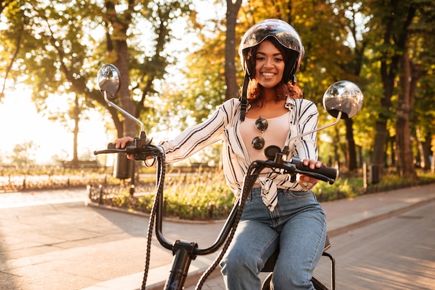 Feliz mulher africana anda de moto moderna no parque e olhando para a câmera