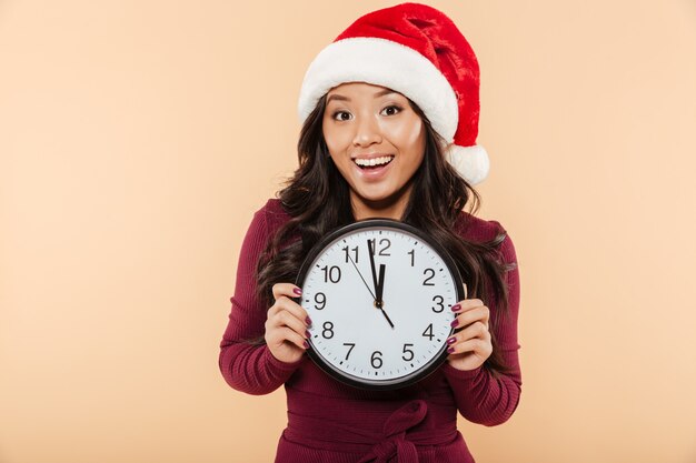Feliz mulher asiática no chapéu de Papai Noel vermelho segurando o relógio mostrando quase 12 comemorando a véspera de ano novo sobre fundo pêssego