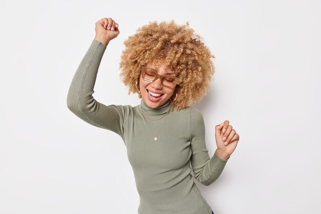 Feliz mulher de cabelos cacheados despreocupada se move ativamente faz a dança do triunfo celebra algo sorri amplamente usa óculos e gola rulê casual isolada sobre fundo branco tem humor otimista