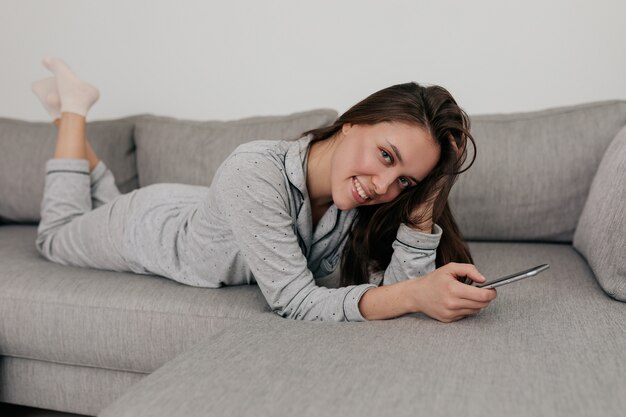 Feliz mulher europeia com um sorriso adorável e cabelo escuro de pijama deitado na carruagem e sorrindo. Retrato interno em casa em dia de sol