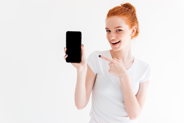 Feliz mulher gengibre mostrando a tela do smartphone em branco e apontando para ele