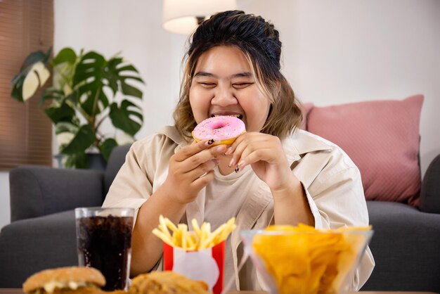 Feliz mulher gorda asiática gosta de comer uma deliciosa rosquinha doce e comida rápida na sala de estar