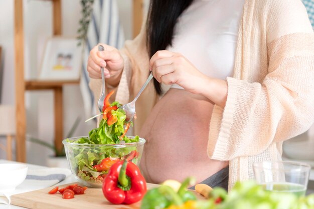 Feliz mulher grávida asiática cozinhando salada em casa fazendo salada verde fresca comendo muitos vegetais diferentes durante a gravidez conceito de gravidez saudável