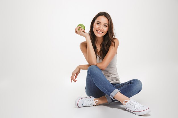 Feliz mulher morena sentada no chão com a apple e olhando para a câmera sobre cinza