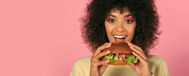 feliz mulher negra com cabelos ondulados, comendo saboroso cheeseburguer em rosa.