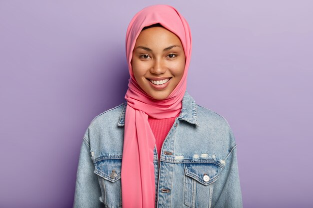 feliz mulher oriental tem religião islâmica, cabeça coberta com véu rosa, sorri suavemente, mostra dentes brancos, isolada contra parede violeta expressa sentimentos e emoções positivas. Etnia