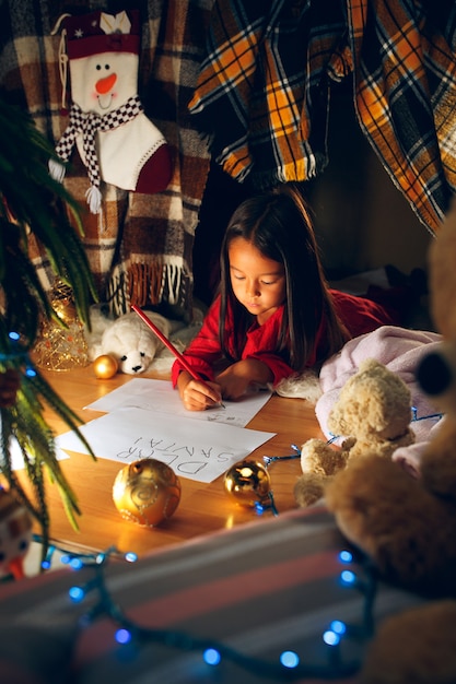 Feliz Natal e Boas Festas. Uma menina linda criança escreve a carta para o Papai Noel perto da árvore de Natal