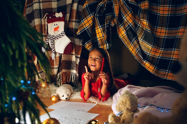 Feliz Natal e Boas Festas. Uma menina linda criança escreve a carta para o Papai Noel perto da árvore de Natal