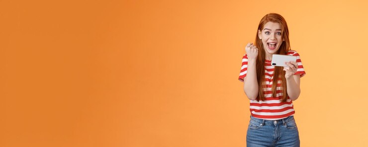 Foto grátis feliz nível de passagem feminina ruiva animada como um incrível gol de pontuação de jogo, segure o smartphone horizontalmente fi