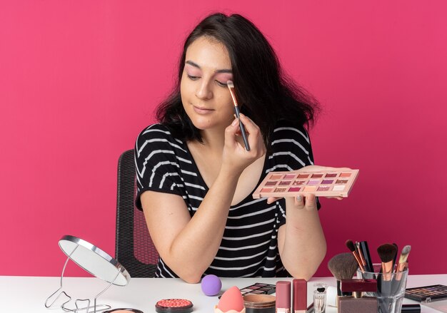 Feliz olhando para o espelho, uma jovem linda se senta à mesa com ferramentas de maquiagem, aplicando a sombra com pincel de maquiagem isolado na parede rosa