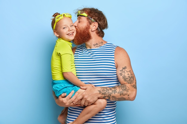 Foto grátis feliz pai e filha usam óculos e roupas de verão, divirtam-se juntos durante o descanso. pai afetuoso carrega a menina, beija-a na bochecha, expressa amor. família e conceito de recreação