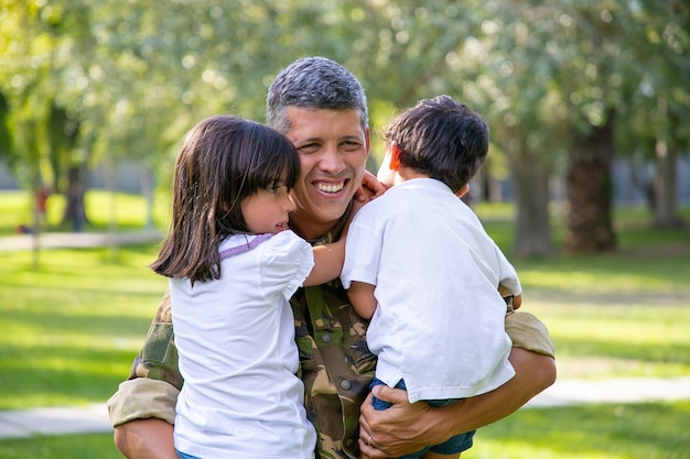 Feliz pai militar encontrando-se com os filhos após a viagem da missão militar, segurando as crianças nos braços e sorrindo. Conceito de reunião familiar ou retorno a casa