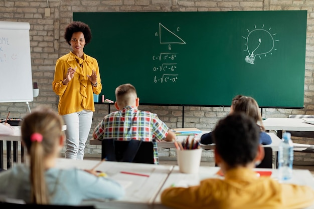 Feliz professora negra falando com seus alunos enquanto dá aula de matemática na sala de aula