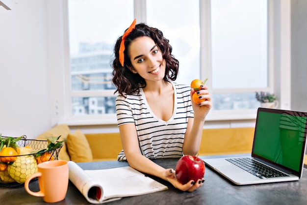 Feliz relaxe o tempo em casa de uma jovem alegre com cabelo cortado e encaracolado, sorrindo na mesa da sala de estar. laptop com tela verde, frutas cítricas, maçã, revista, chá, refrigeração em apartamento moderno