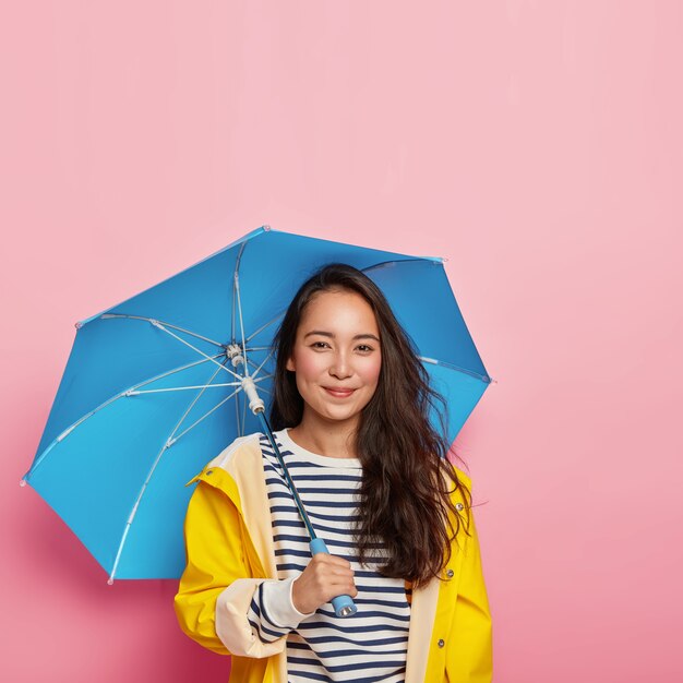 Feliz senhora oriental de cabelos escuros com beleza natural, se sente seca e protegida, usa capa de chuva impermeável, carrega guarda-chuva, aproveita o tempo livre durante o dia chuvoso de outono