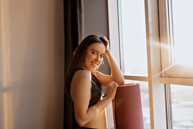 Foto grátis feliz senhora sorridente com penteado escuro de uniforme está segurando o tapete de ioga e olhando para a câmera à luz do sol de manhã em casa