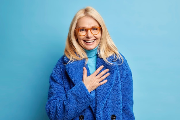 Feliz sênior loira mulher europeia divertida com uma piada humorística, risos positivamente mantém a mão no peito vestida com casaco azul de inverno.