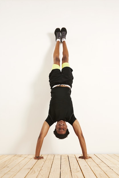 feliz sorridente jovem modelo preto em roupas de ginástica pretas e amarelas, fazendo parada de mãos contra uma parede branca no chão de madeira.