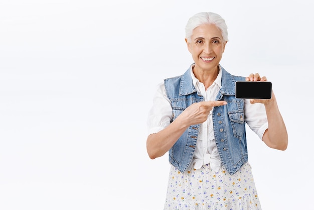 Feliz velhinha moderna com corte de cabelo grisalho penteado segurando o smartphone horizontalmente apontando a tela do celular e sorrindo como dando conselhos que download de aplicativo recomenda página de internet ou promoção