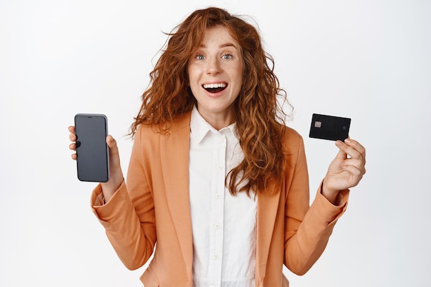Feliz vendedora com cabelo ruivo encaracolado, mostrando a tela do celular e o cartão de crédito sorrindo espantado em pé de terno e camisa de escritório contra fundo branco