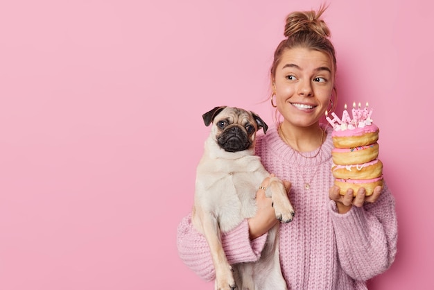 Feliz woma encantada comemora aniversário de poses de animais de estimação favoritas com cachorro e deliciosos donuts com velas usa suéter de malha isolado sobre fundo rosa espaço em branco para seu texto promocional