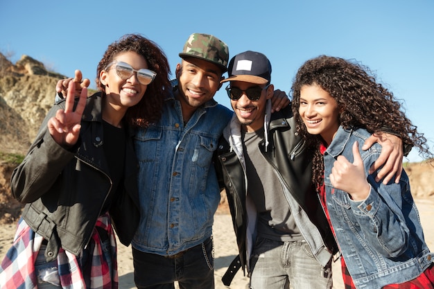 Felizes amigos africanos caminhando ao ar livre na praia, mostrando os polegares