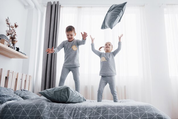 Felizes crianças brincando no quarto branco. Menino e menina, irmão e irmã jogam na cama de pijama. Roupa de dormir e roupa de cama para bebê e criança. Família em casa