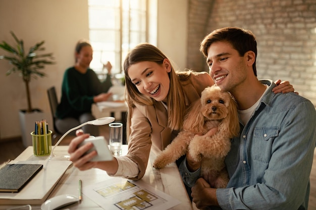 Felizes freelancers usando telefone celular enquanto tiram selfie com um cachorro no escritório