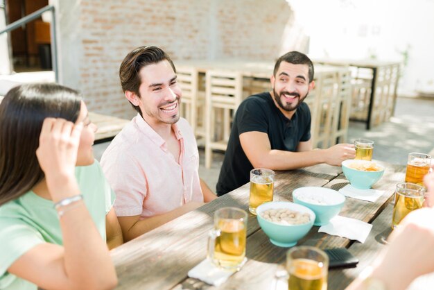 Felizes homens e mulheres jovens se divertindo enquanto estão sentados juntos em um bar ao ar livre. Amigos alegres conversando e bebendo cerveja