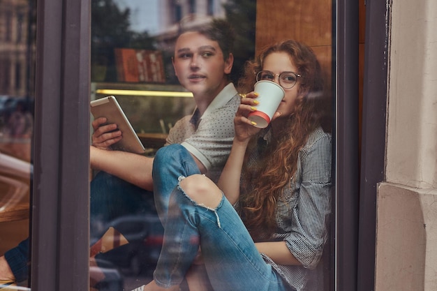 Felizes jovens estudantes bebendo café e usando um tablet digital sentado no parapeito de uma janela em um campus universitário durante um intervalo.