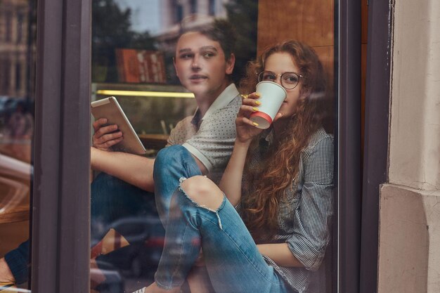 Felizes jovens estudantes bebendo café e usando um tablet digital sentado no parapeito de uma janela em um campus universitário durante um intervalo.