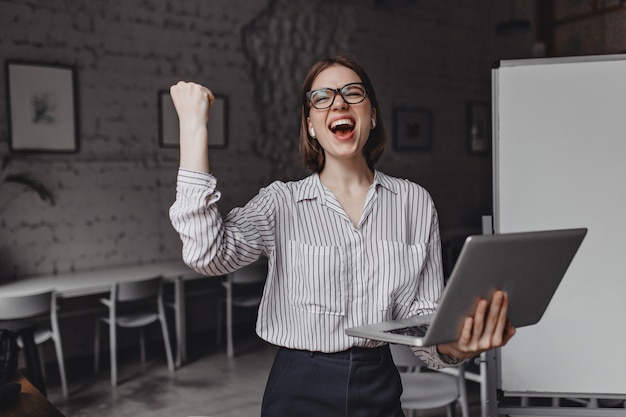 Foto grátis felizmente, a menina grita e faz o gesto com a mão vencedor, segurando o laptop e posando no escritório contra o fundo do quadro.