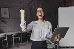 Foto grátis felizmente, a menina grita e faz o gesto com a mão vencedor, segurando o laptop e posando no escritório contra o fundo do quadro.