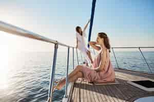 Foto grátis fêmea adulta tenra e atraente, passando um tempo no barco. mulher fica na proa do iate com olhar sonhador, enquanto a amiga se senta ao lado, ambos se sentindo no paraíso