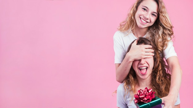 Fêmea cobrindo os olhos da amiga dando caixa de presente contra um fundo rosa