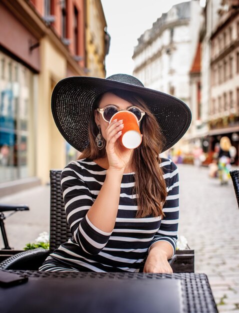 Fêmea de chapéu de verão e vestido com listras tomando café no café de verão.