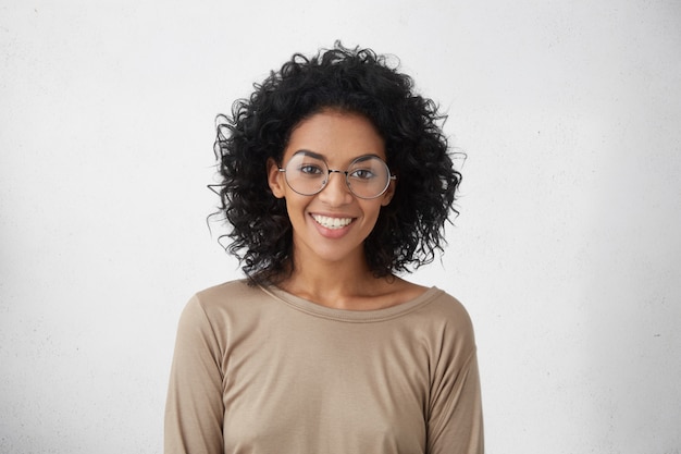 Foto grátis fêmea de raça mista despreocupada e relaxada, muito jovem, usando grandes óculos redondos, sorrindo amplamente, sentindo-se entusiasmada por passar férias no exterior