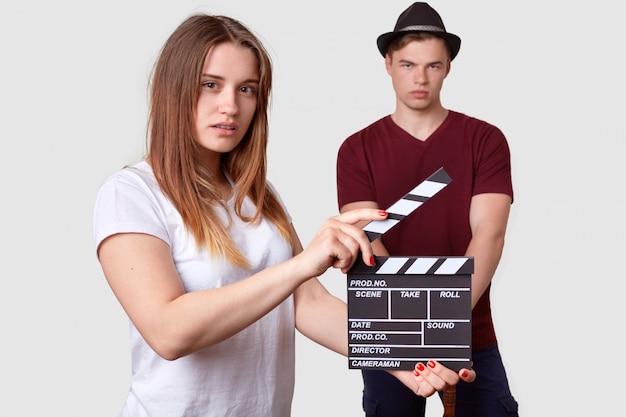 Foto grátis fêmea em camiseta branca possui claquete, filma cena, homem estiloso sério fica em primeiro plano, usa arnês elegante et camiseta, envolvidos na produção do filme. conceito de filme