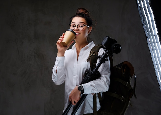 Fêmea viajante detém câmera fotográfica digital e café para viagem sobre fundo cinza.