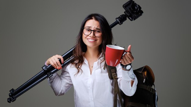 Fêmea viajante detém câmera fotográfica digital e uma xícara de café vermelha sobre fundo cinza.